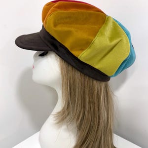 May include: A multi-colored newsboy cap featuring panels of red, orange, yellow, lime green, and blue velvet fabric. The cap has a dark brown brim and a matching dark brown band that encircles the base, providing a structured fit for the head.