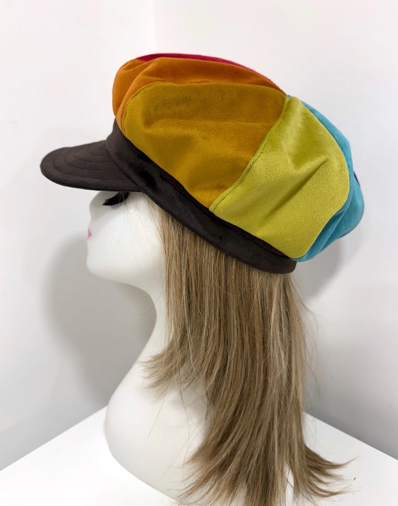 May include: A multi-colored newsboy cap featuring panels of red, orange, yellow, lime green, and blue velvet fabric. The cap has a dark brown brim and a matching dark brown band that encircles the base, providing a structured fit for the head.