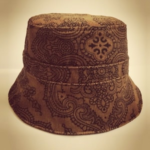 May include: A brown corduroy bucket hat with a dark brown paisley design. The hat has a structured brim and a band around the crown. The fabric has a ribbed texture.