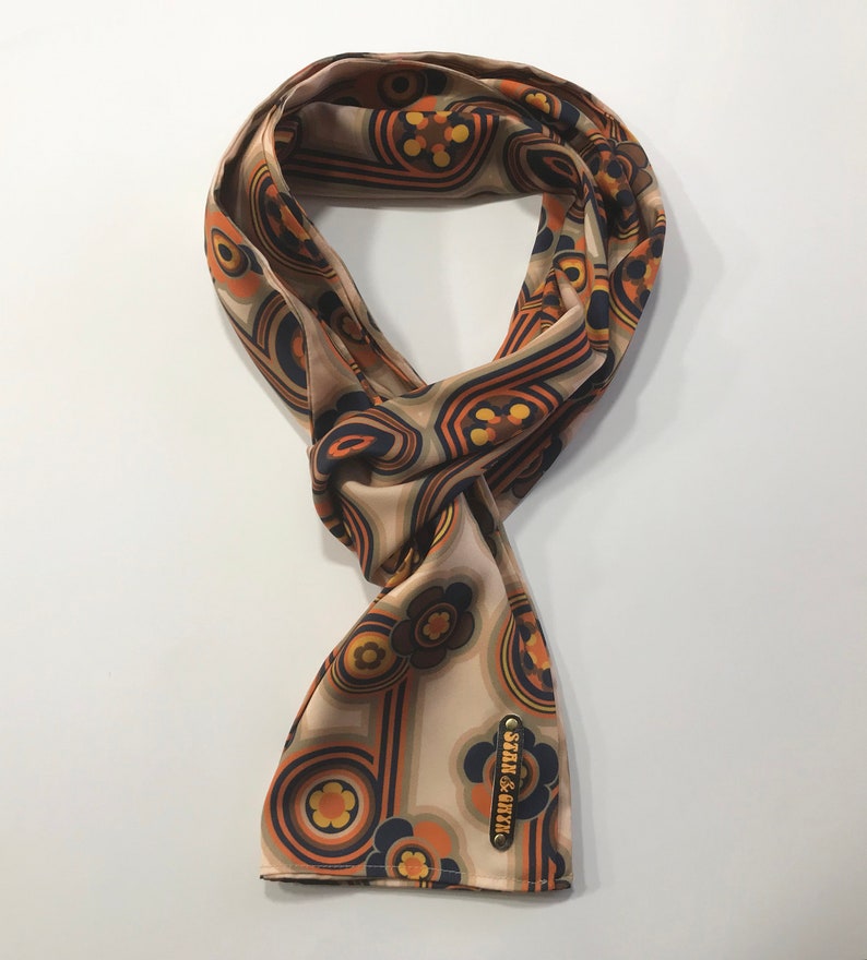 Pu&ograve; includere: Un foulard di seta beige con un motivo floreale e geometrico retr&ograve; in tonalit&agrave; arancio, marrone e blu. Il foulard ha una piccola etichetta dorata con il testo "Stark & River".