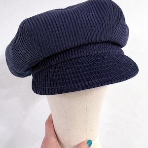 May include: A navy blue corduroy newsboy cap featuring a structured crown with vertical ribbed texture and a short, stiff brim. The hat is displayed on a neutral-colored mannequin head form, showcasing the fabric's deep blue color and texture.