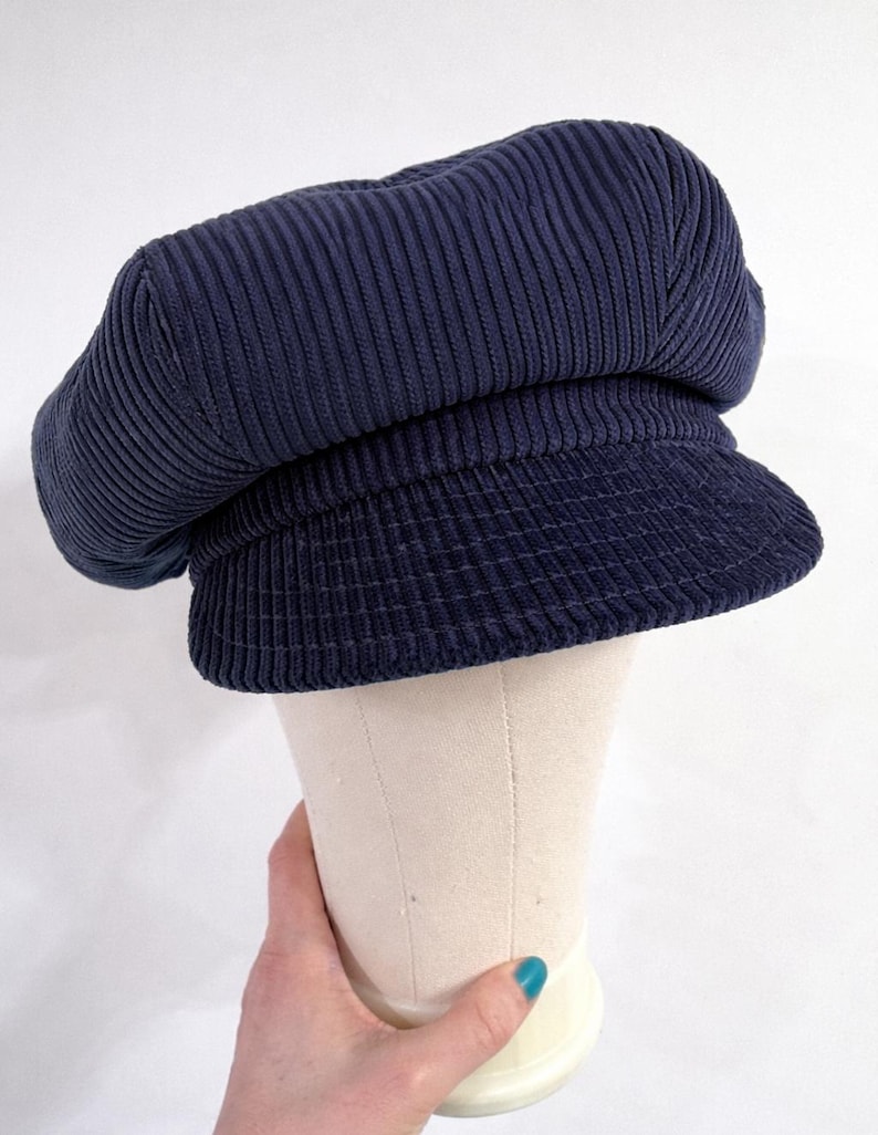 May include: A navy blue corduroy newsboy cap featuring a structured crown with vertical ribbed texture and a short, stiff brim. The hat is displayed on a neutral-colored mannequin head form, showcasing the fabric's deep blue color and texture.
