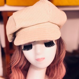 May include: A beige corduroy newsboy cap with a structured crown and a short, stiff brim. The hat features a textured, ribbed fabric surface and is displayed on a mannequin head with long, wavy pink hair and prominent black false eyelashes.
