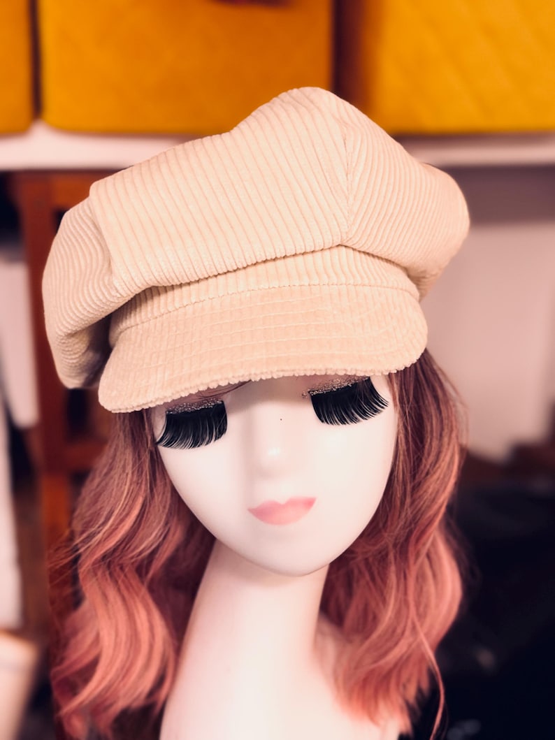 May include: A beige corduroy newsboy cap with a structured crown and a short, stiff brim. The hat features a textured, ribbed fabric surface and is displayed on a mannequin head with long, wavy pink hair and prominent black false eyelashes.
