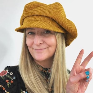 May include: A mustard yellow corduroy newsboy cap featuring a structured brim and a soft, multi-paneled crown. The fabric has a distinct vertical ribbed texture throughout, providing a classic look with a rounded shape that sits comfortably on the head.