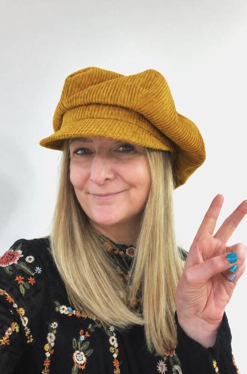 May include: A mustard yellow corduroy newsboy cap featuring a structured brim and a soft, multi-paneled crown. The fabric has a distinct vertical ribbed texture throughout, providing a classic look with a rounded shape that sits comfortably on the head.