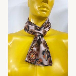 Pu&ograve; includere: Un foulard di seta con motivo marrone, beige, arancione e blu con un'etichetta in metallo dorato che recita "Stan & Anya".