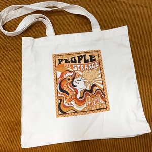 May include: White canvas tote bag featuring a retro-style graphic of a woman with long, wavy hair and the text "People are strange, Faces come out of the rain".