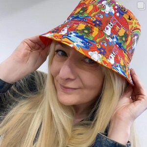 May include: A colorful bucket hat with a psychedelic pattern featuring mushrooms, rabbits, and flowers. The hat is orange, yellow, red, blue, and green.