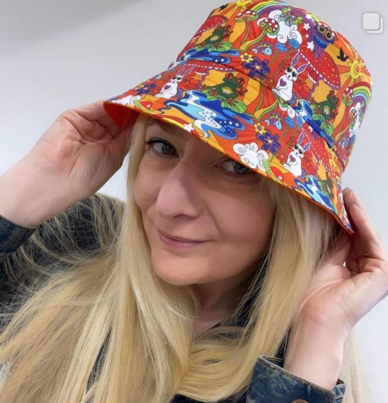 May include: A colorful bucket hat with a psychedelic pattern featuring mushrooms, rabbits, and flowers. The hat is orange, yellow, red, blue, and green.