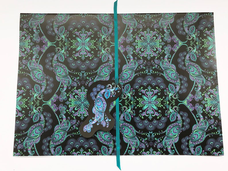 K&ouml;nnte beinhalten: Schwarzes Geschenkpapier mit einem t&uuml;rkisfarbenen Paisley-Muster und einem Pfauen-Design. Das Papier ist mit einem t&uuml;rkisfarbenen Band gebunden.