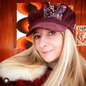 May include: A woman with long blonde hair is wearing a burgundy corduroy newsboy cap with a floral patterned fabric panel. The cap has a wide brim and a slightly curved crown.