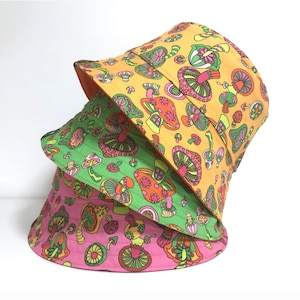 May include: Three bucket hats stacked on top of each other. The top hat is yellow with a mushroom print. The middle hat is green with a mushroom print. The bottom hat is pink with a mushroom print.