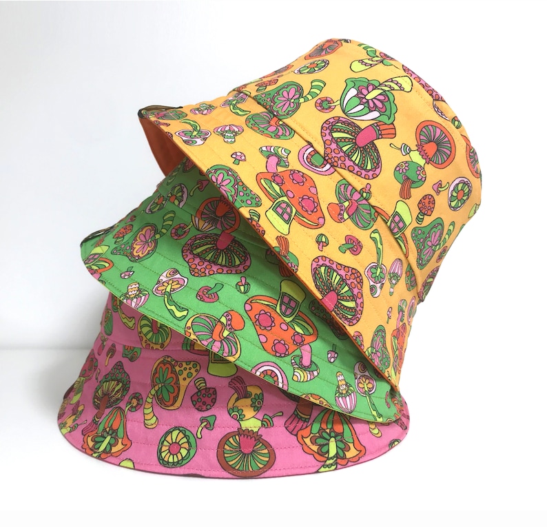 May include: Three bucket hats stacked on top of each other. The top hat is yellow with a mushroom print. The middle hat is green with a mushroom print. The bottom hat is pink with a mushroom print.