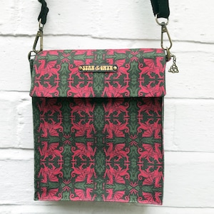 May include: A green and red canvas crossbody bag with a dragon pattern. The bag has a flap closure and a metal clasp. The bag has a metal tag with the text "Stan & Gwyn" on it.