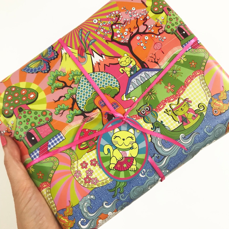 May include: Pink and green wrapping paper with a whimsical design featuring mushrooms, flowers, and cats. The paper is tied with a pink ribbon and a gift tag with a cartoon cat.