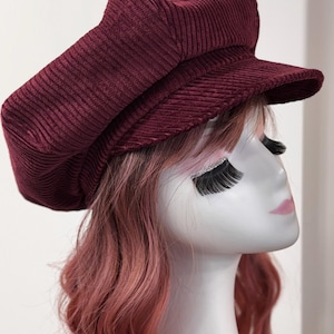 May include: A deep burgundy corduroy newsboy cap featuring a structured crown and a short, stiff brim. The hat is crafted from a textured, ribbed fabric with a classic multi-paneled design, providing a rounded silhouette that sits atop the head.