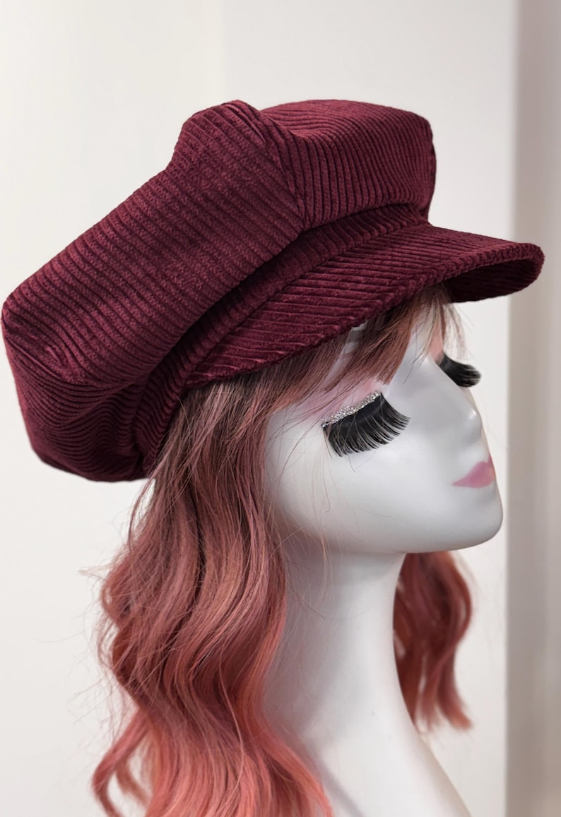 May include: A deep burgundy corduroy newsboy cap featuring a structured crown and a short, stiff brim. The hat is crafted from a textured, ribbed fabric with a classic multi-paneled design, providing a rounded silhouette that sits atop the head.