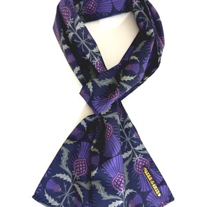 May include: A navy blue silk scarf with a purple thistle pattern. The scarf is tied in a knot and the brand name "Satin & Silk" is visible on the bottom right corner.