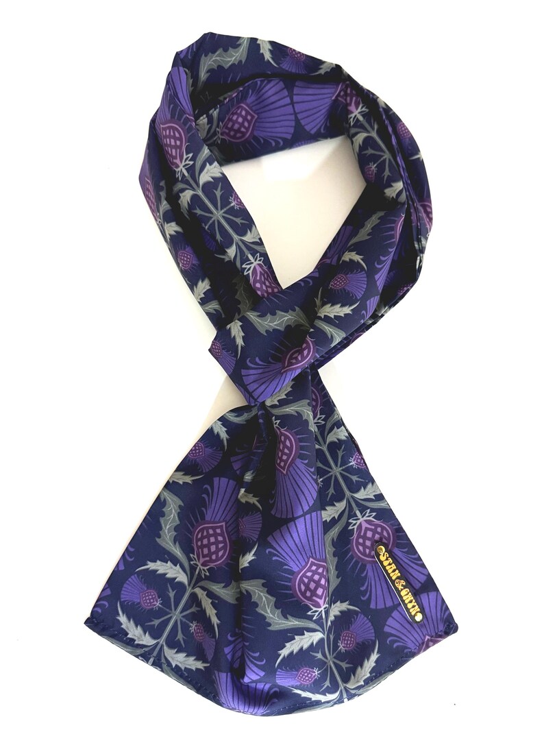 May include: A navy blue silk scarf with a purple thistle pattern. The scarf is tied in a knot and the brand name "Satin & Silk" is visible on the bottom right corner.