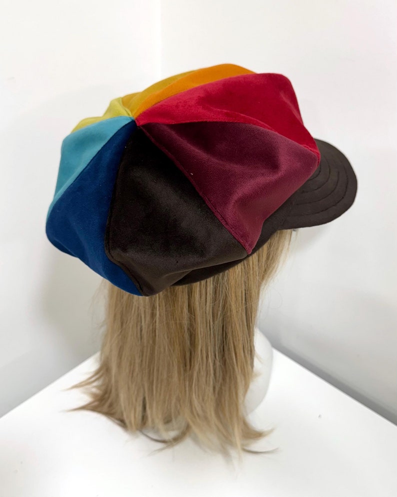 May include: A multi-colored newsboy cap featuring panels of velvet fabric in shades of red, orange, yellow, light blue, dark blue, and dark brown. The cap has a structured dark brown brim with visible stitching and a soft, rounded crown shape.