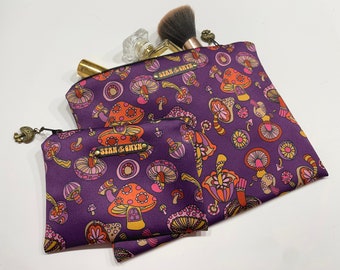 May include: Two purple zippered makeup bags with a mushroom and flower pattern. The larger bag has the text "Sean & Gwyn" on it. The smaller bag has the text "Sean & Gwyn" on it.