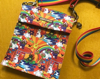 May include: A rectangular crossbody bag featuring a vibrant, multi-coloured print of rabbits, owls, mushrooms, rainbows, and planets on an orange background. The bag has a flap with a metal tag reading STAN & GYNX and a rainbow-striped adjustable strap.