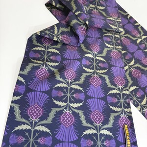 May include: A purple and green patterned silk scarf with a repeating thistle design. The scarf has a navy blue background and the text "OSCAR & ALEX" is printed on the bottom right corner.