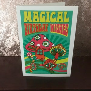 Op de afbeelding: Een groene kaart met de tekst "Magical Birthday Wishes" in gele en rode letters. De kaart heeft een cartoonillustratie van kleurrijke paddenstoelen met witte stippen.