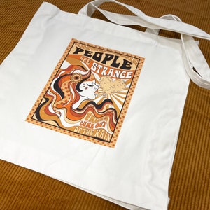 May include: White canvas tote bag with a retro-style graphic design featuring a woman with long, flowing hair and the text "People are strange, Faces came out of the rain".