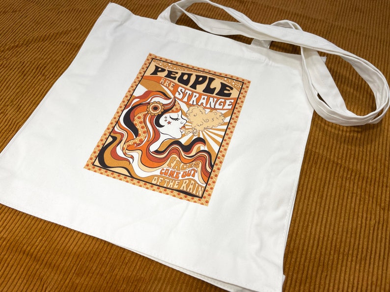 May include: White canvas tote bag with a retro-style graphic design featuring a woman with long, flowing hair and the text "People are strange, Faces came out of the rain".