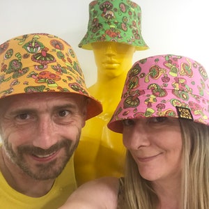 May include: Two people wearing colorful bucket hats with mushroom designs. The hats are yellow, green, and pink with a variety of mushroom illustrations. The hats have a wide brim and are made of a soft, comfortable fabric.