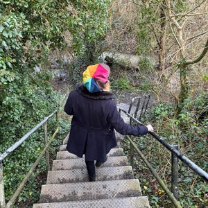 May include: A multi-colored beret featuring segments of yellow, orange, red, pink, blue, and brown. The beret is worn with a dark navy blue coat that has a dark faux fur collar, paired with black trousers, while descending a set of metal stairs.
