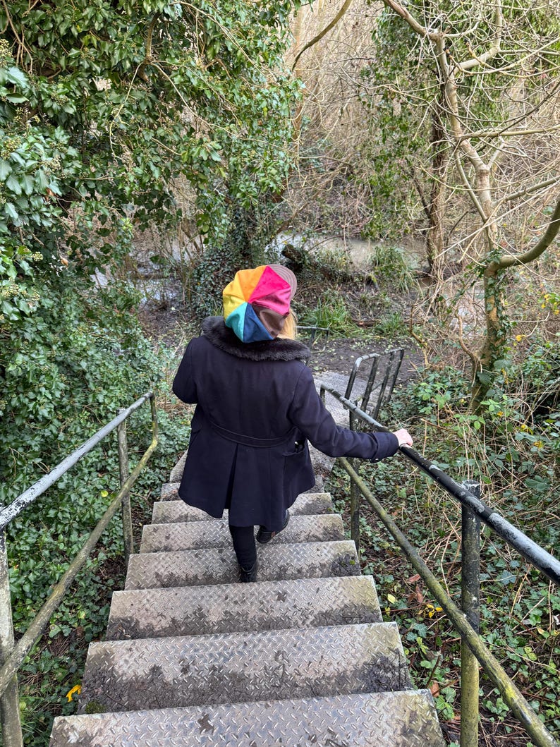 May include: A multi-colored beret featuring segments of yellow, orange, red, pink, blue, and brown. The beret is worn with a dark navy blue coat that has a dark faux fur collar, paired with black trousers, while descending a set of metal stairs.