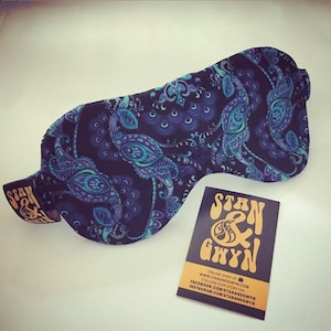 Könnte beinhalten: Eine schwarze Schlafmaske mit Paisley-Muster in Blautönen und Lila. Die Maske ist aus weichem Stoff gefertigt und hat ein elastisches Band. Die Maske wird mit einer Visitenkarte gezeigt, auf der "Stan & Gwyn" steht, mit Kontaktinformationen für ihren Online-Shop.