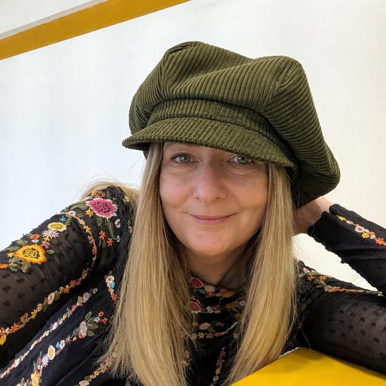 May include: An olive green corduroy newsboy cap with a structured brim and a soft, rounded crown. The fabric features a distinct vertical ribbed texture. The cap is worn, showcasing its full shape, panel construction, and deep, earthy color tone.
