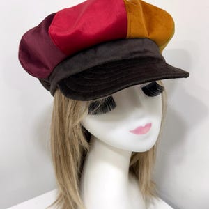 May include: A multi-colored newsboy cap featuring panels in shades of deep burgundy, bright red, and mustard yellow. The hat includes a dark brown brim and a matching dark brown band, constructed from a soft, smooth fabric with a structured shape.