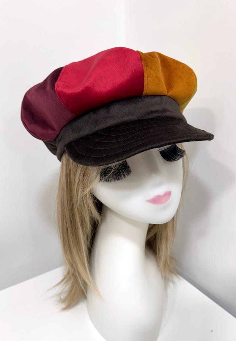 May include: A multi-colored newsboy cap featuring panels in shades of deep burgundy, bright red, and mustard yellow. The hat includes a dark brown brim and a matching dark brown band, constructed from a soft, smooth fabric with a structured shape.