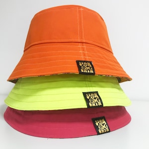 May include: Three bucket hats stacked on top of each other. The top hat is orange, the middle hat is neon green, and the bottom hat is pink. Each hat has a black patch with gold lettering that says "Stan & Bryn".