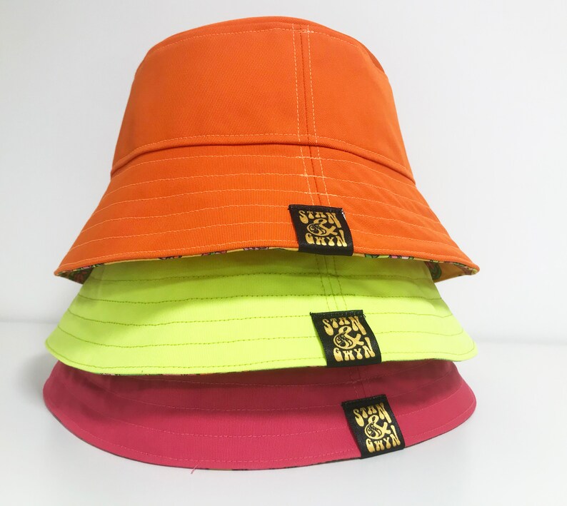 May include: Three bucket hats stacked on top of each other. The top hat is orange, the middle hat is neon green, and the bottom hat is pink. Each hat has a black patch with gold lettering that says "Stan & Bryn".