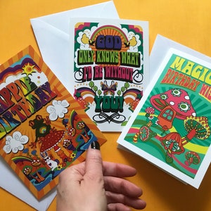 Op de afbeelding: Drie kleurrijke wenskaarten met psychedelische ontwerpen. De eerste kaart heeft een regenboogachtergrond met de tekst "Happy Birthday" in een groovy lettertype. De tweede kaart heeft een groene achtergrond met de tekst "God Only Knows What I'd Be Without You!" in een psychedelisch lettertype. De derde kaart heeft een groene achtergrond met de tekst "Magical Birthday Wishes" in een psychedelisch lettertype.
