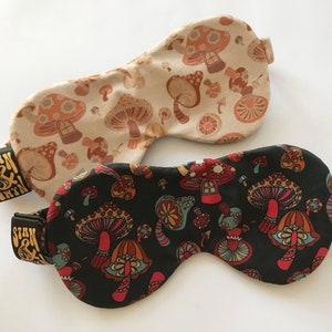 May include: Two sleep masks with a mushroom print. One mask is beige with brown mushrooms and the other is black with colourful mushrooms. Both masks have a gold label that says "Stan & Gryn."