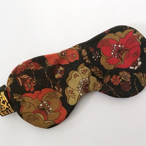 Könnte beinhalten: Eine schwarze Schlafmaske mit einem floralen Muster in Rot-, Orange- und Brauntönen. Die Maske hat ein elastisches Band mit dem aufgedruckten Text "Stan & Gryn".