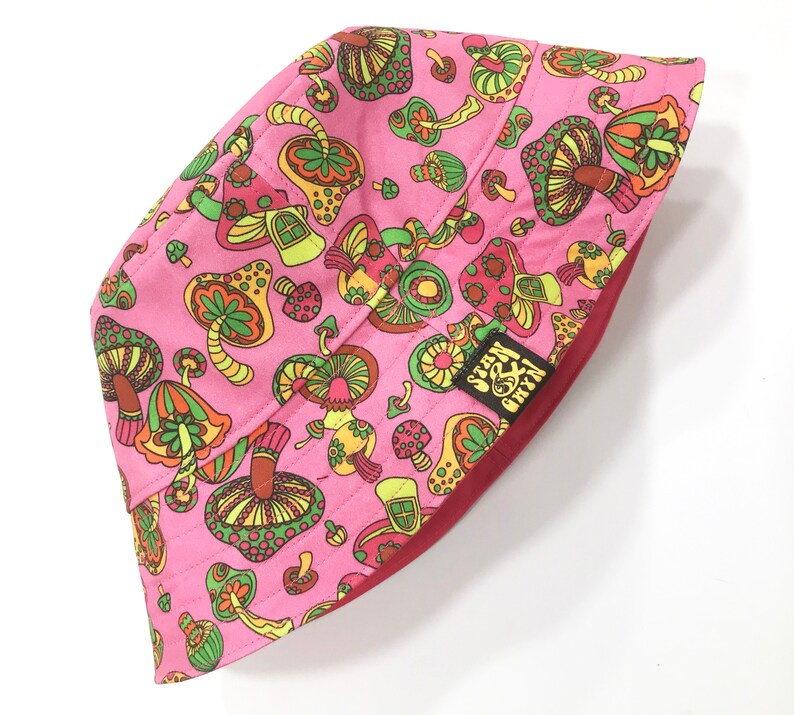 May include: Pink bucket hat with a mushroom and flower pattern. The hat has a red underside and a small green and yellow patch with the text "Green & Grey" on it.