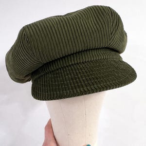 May include: An olive green corduroy newsboy cap featuring a structured, rounded crown and a short, stiff brim. The fabric has a distinct vertical ribbed texture throughout the entire surface of the hat, providing a consistent look and feel.