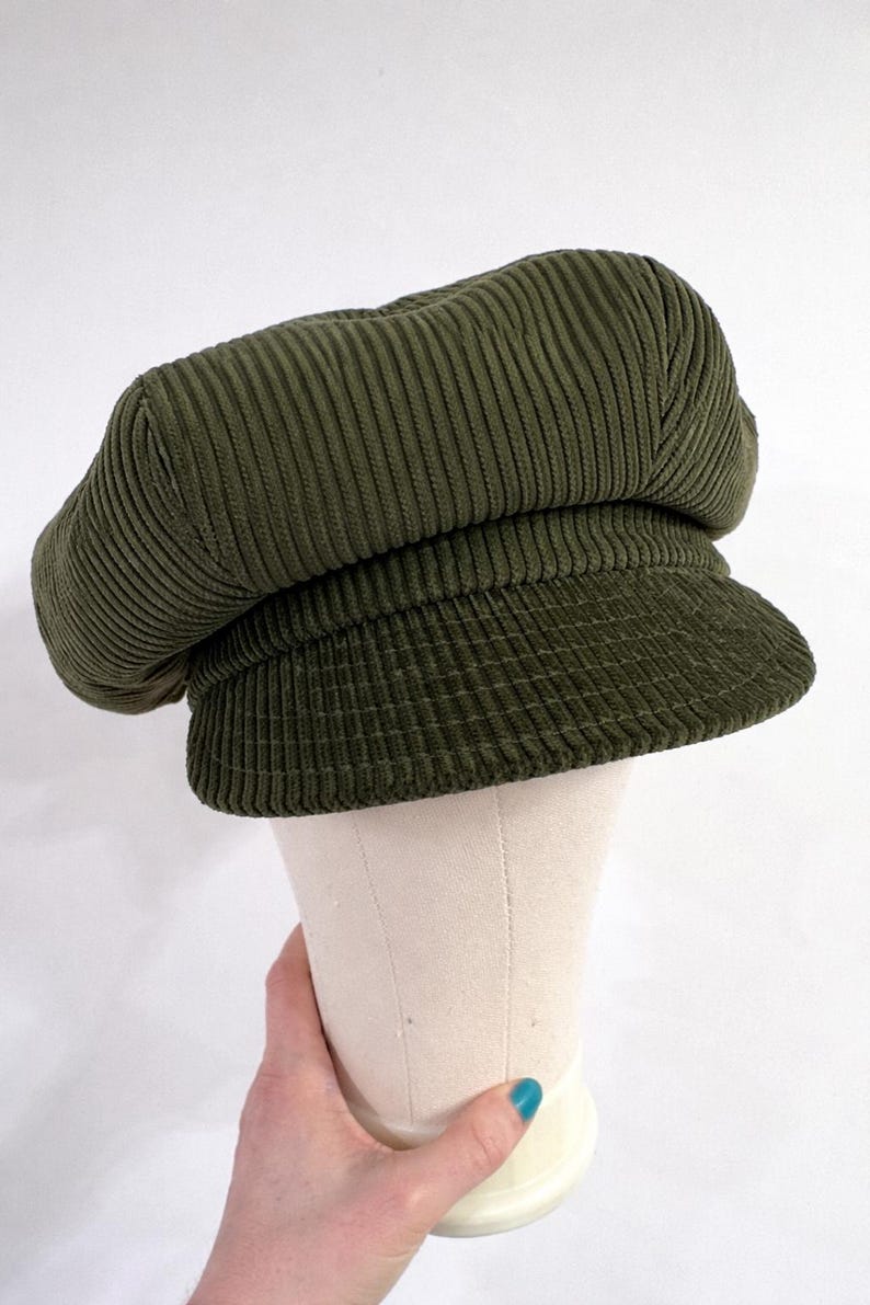 May include: An olive green corduroy newsboy cap featuring a structured, rounded crown and a short, stiff brim. The fabric has a distinct vertical ribbed texture throughout the entire surface of the hat, providing a consistent look and feel.