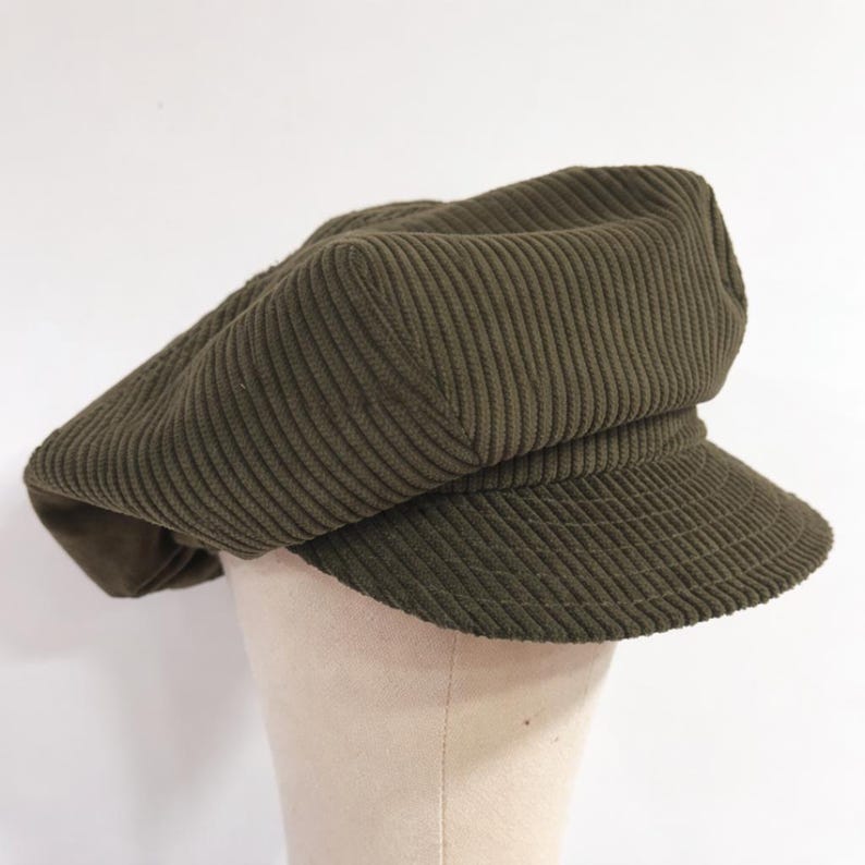 May include: An olive green corduroy newsboy cap featuring a structured crown and a short, stiff brim. The fabric has a distinct vertical ribbed texture throughout the entire surface of the hat, providing a consistent look from the crown to the visor.