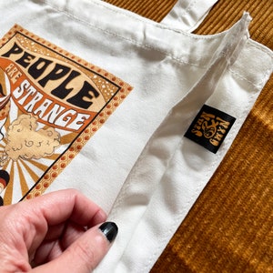 May include: White canvas tote bag with a retro graphic design featuring a woman with a sunflower and the text "People are strange". The bag has a black label with the text "Seven & Chix" on it.