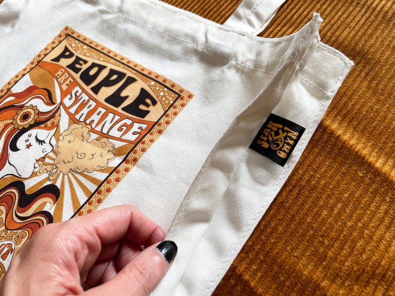 May include: White canvas tote bag with a retro graphic design featuring a woman with a sunflower and the text "People are strange". The bag has a black label with the text "Seven & Chix" on it.