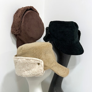 May include: Three corduroy hats with ear flaps. The hats are brown, black, and beige. The beige hat has a white fleece lining.